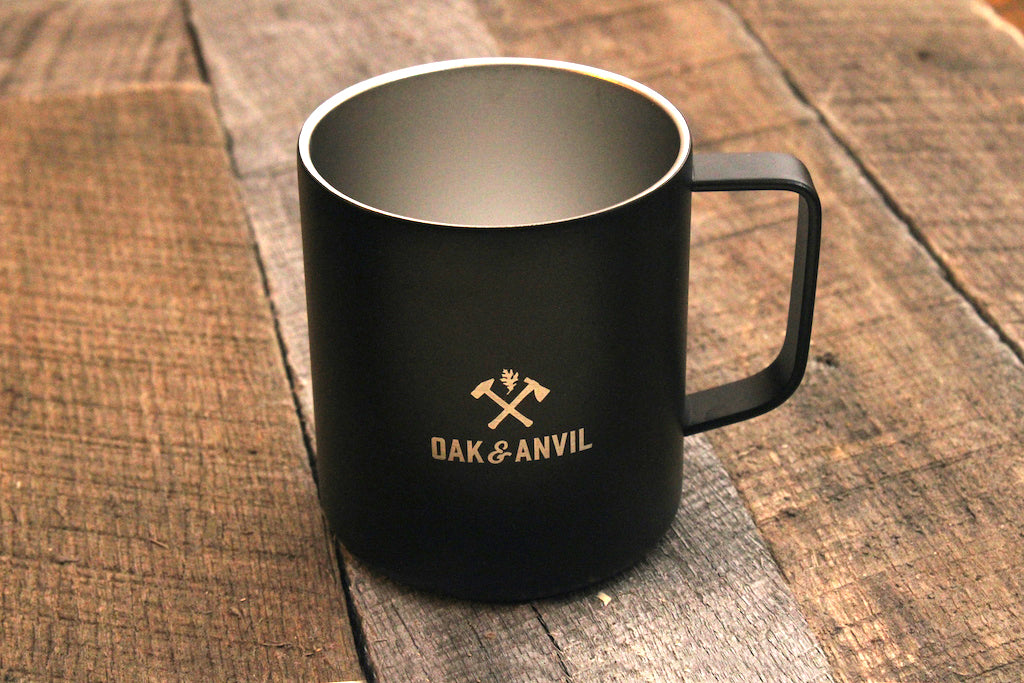 Black stainless steel insulated mug with Oak and Anvil logo on grey wood